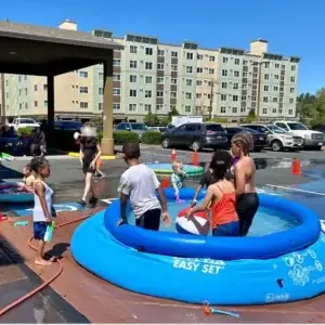 Diverse children play in a blue inflatable pool outside apartment buildings on a sunny day.