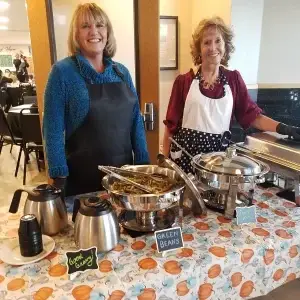 Two smiling women serve fall-themed buffet food: green beans, gravy, sweet potatoes.