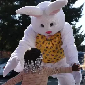A child with beaded braids reaches for a white Easter Bunny in a yellow vest.