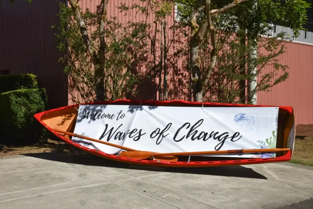 Bright red rowboat holds a banner reading "Welcome to Waves of Change" with oars.