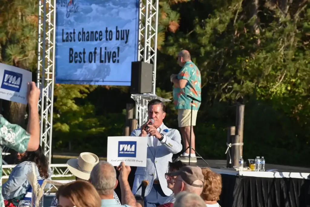 Man speaks on stage, audience holds signs. Screen: 'Last chance to buy Best of Live!'.