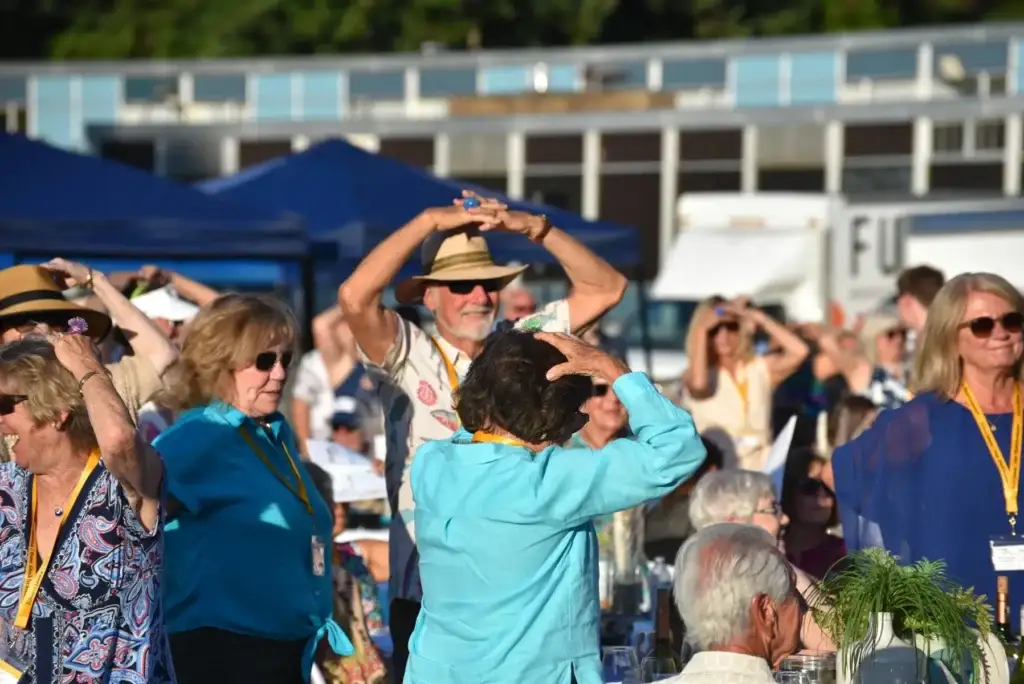 Lively group of older adults dancing outdoors, many raising hands over heads, smiling.