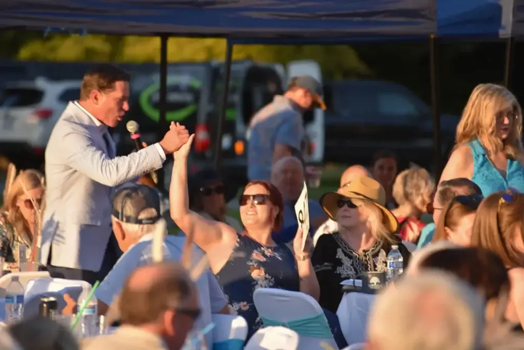 Auctioneer high-fives smiling woman holding bid paddle at lively outdoor event.