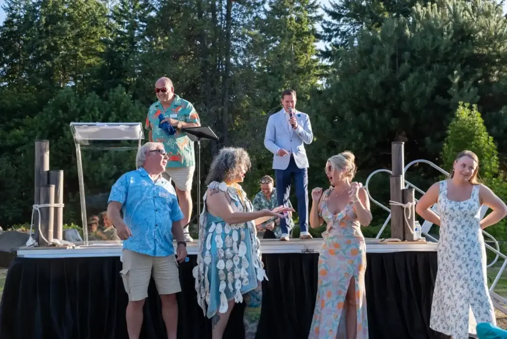 Outdoor stage event. Man speaks, woman wears oyster shell dress, others watch and react.