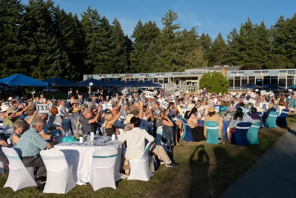 Many people raise numbered paddles at a lively outdoor auction on a sunny grassy field.