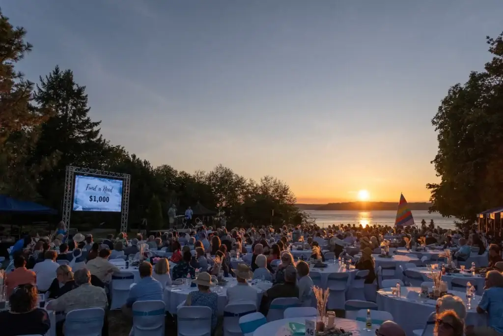 Outdoor fundraiser by lake at sunset. Guests at tables, screen shows 'Fund a Need'.