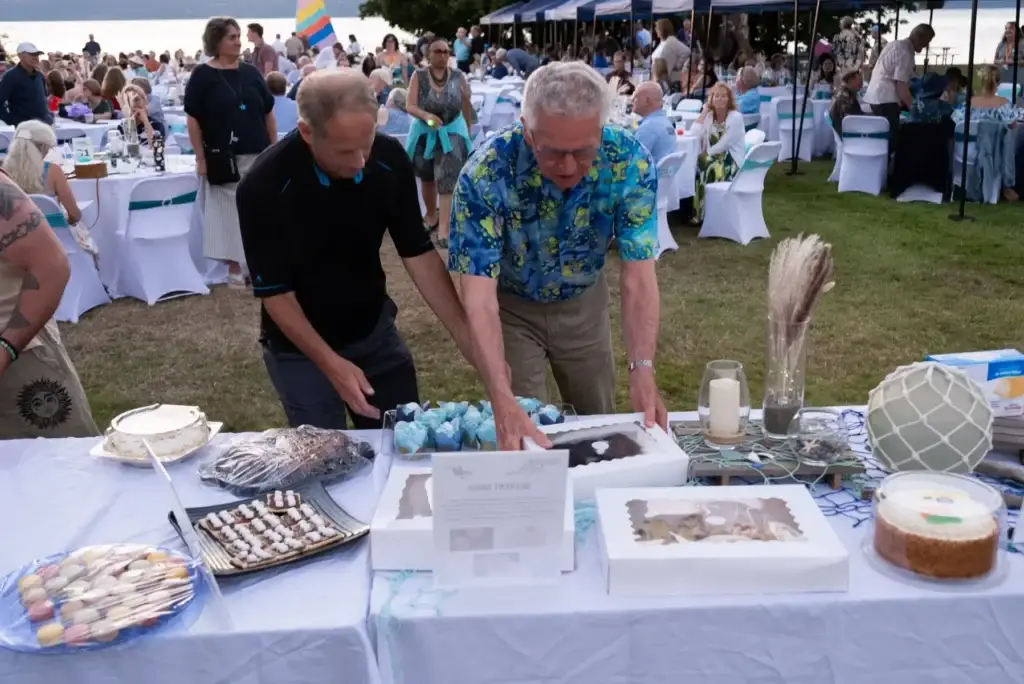 Two men arrange cakes and blue cupcakes on a nautical-themed dessert table outdoors.