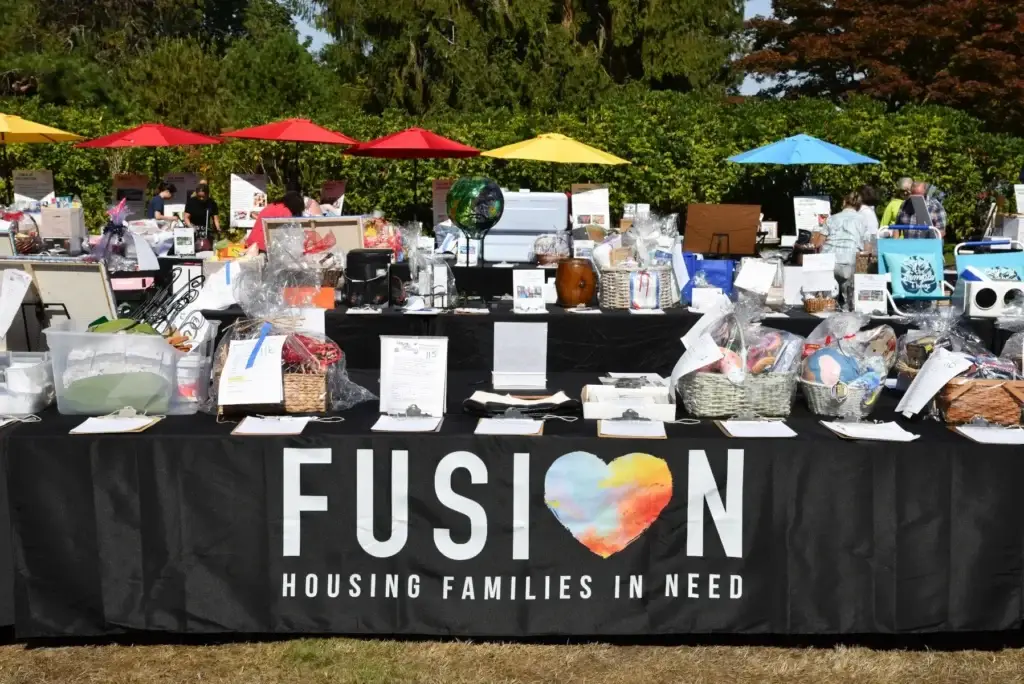 Outdoor fundraiser: FUSION banner on table packed with gift baskets under colorful umbrellas.