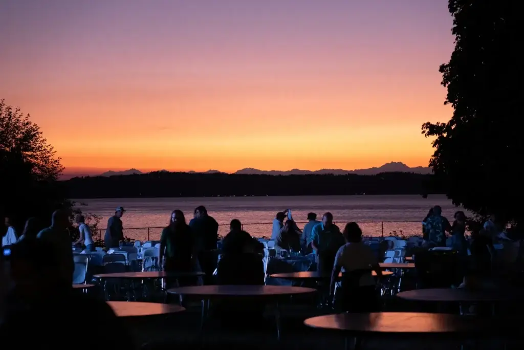 Vibrant purple and orange sunset over water and mountains, with silhouetted people at tables.