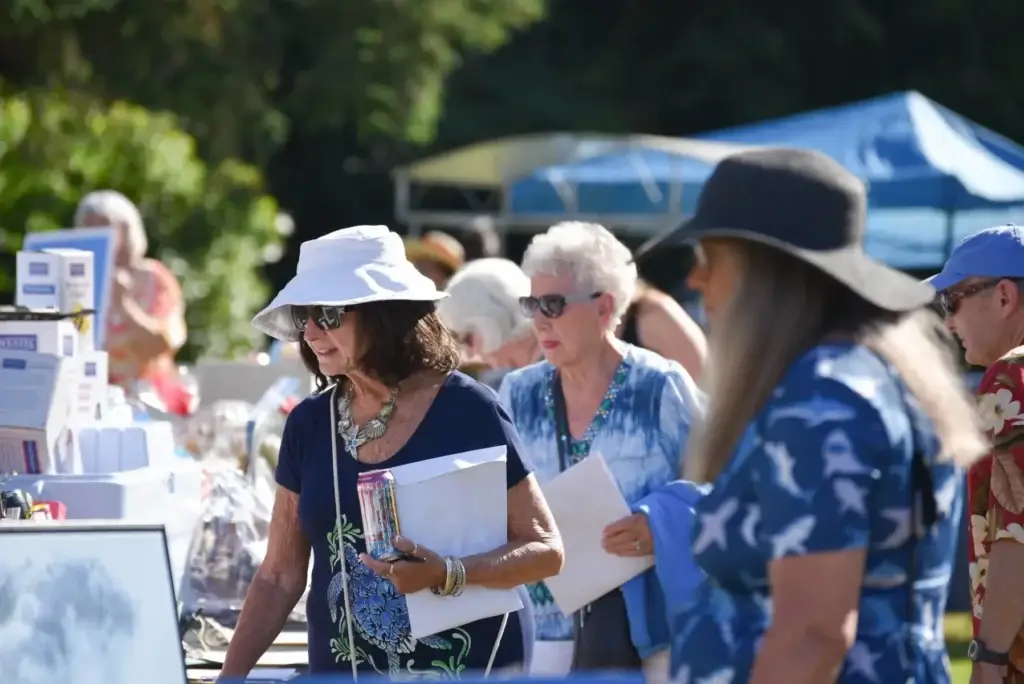 People in sun hats and casual clothes browse an outdoor market on a sunny day.