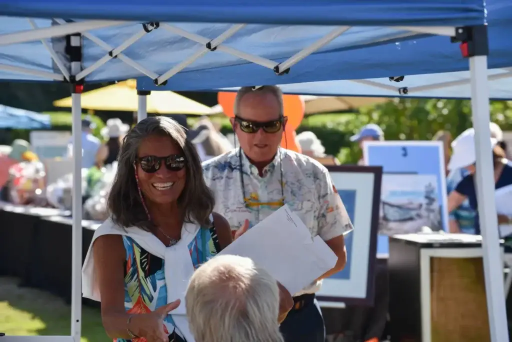 Smiling woman in sunglasses, tropical dress, under blue tent at an outdoor event.