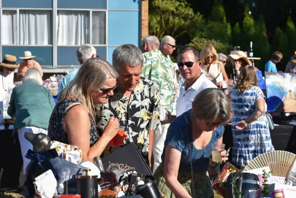 Adults browse items on tables at a sunny outdoor market or gathering.