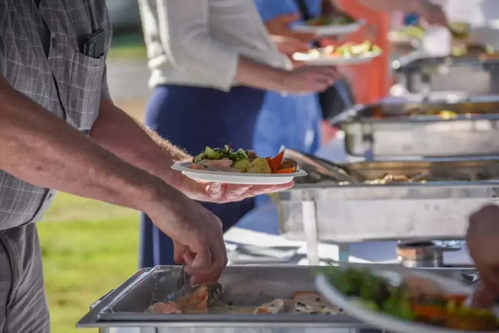Hands serve diverse food from metal chafing dishes at a busy outdoor buffet.