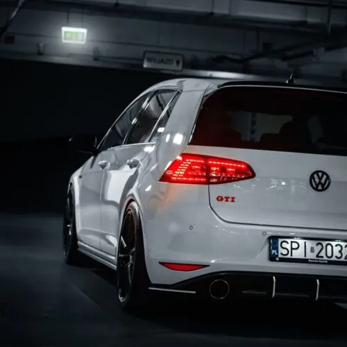 Ein weißer Volkswagen Golf GTI, der als Projektbotschafter dient, stellt seine beleuchteten Rückleuchten in einer schwach beleuchteten Garage zur Schau. Das Auto wird von der hinteren linken Seite betrachtet, wodurch sein sportliches Design und ein Teil des Nummernschilds hervorgehoben werden.