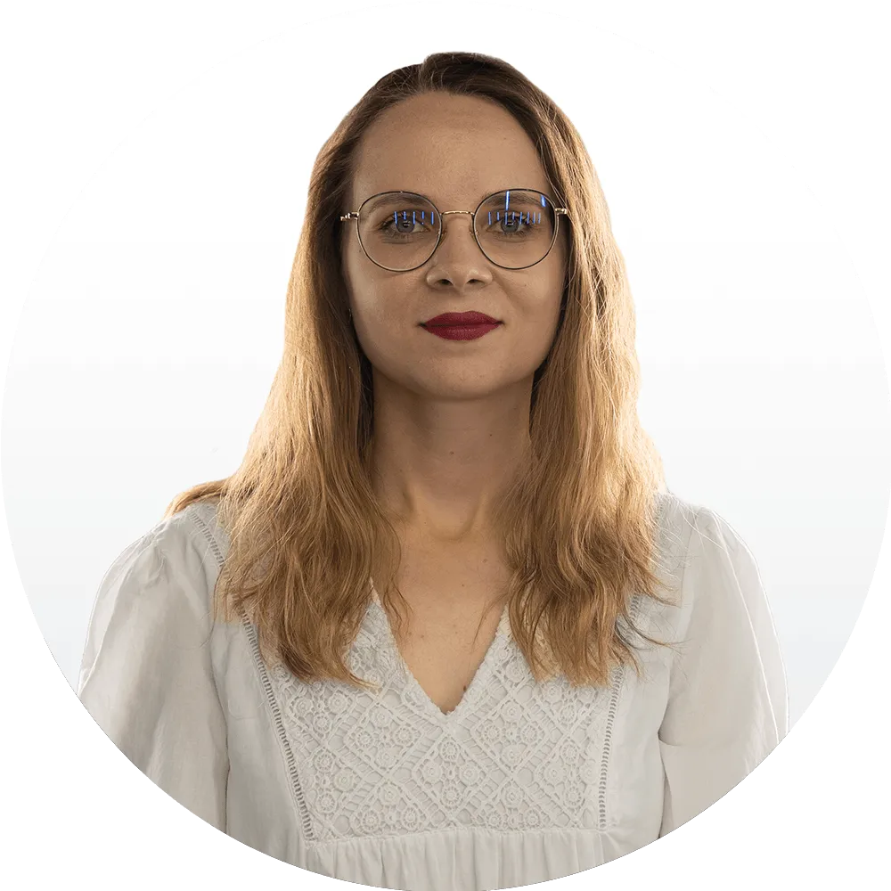 Jagoda Cieślak A woman with long, wavy light brown hair, wearing round glasses and a white blouse with lace details, stands in front of a plain white background, looking directly at the camera.
