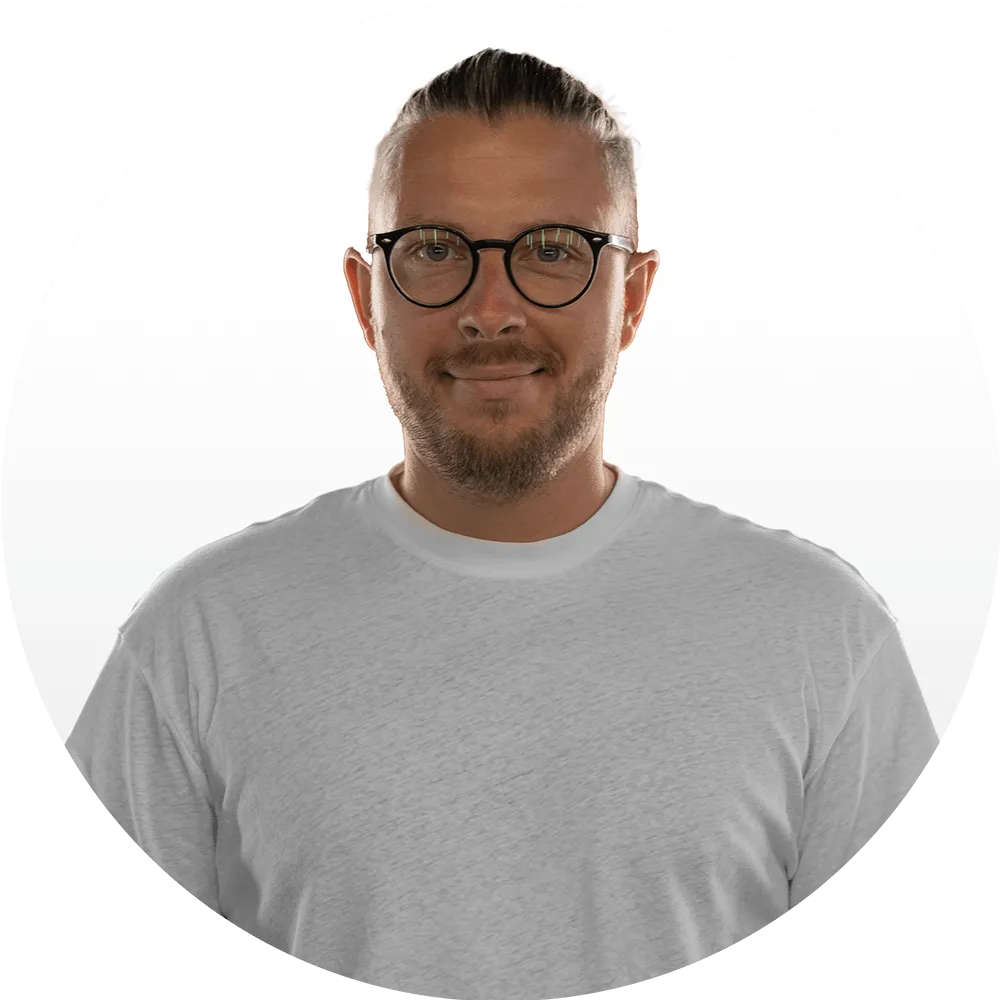 Michał Styperek A man with short, slicked-back hair, glasses, and a trimmed beard is wearing a plain white t-shirt and smiling gently at the camera. Meet Us—the Good Stuff team brings warmth and authenticity to our About Us page. The background is white.