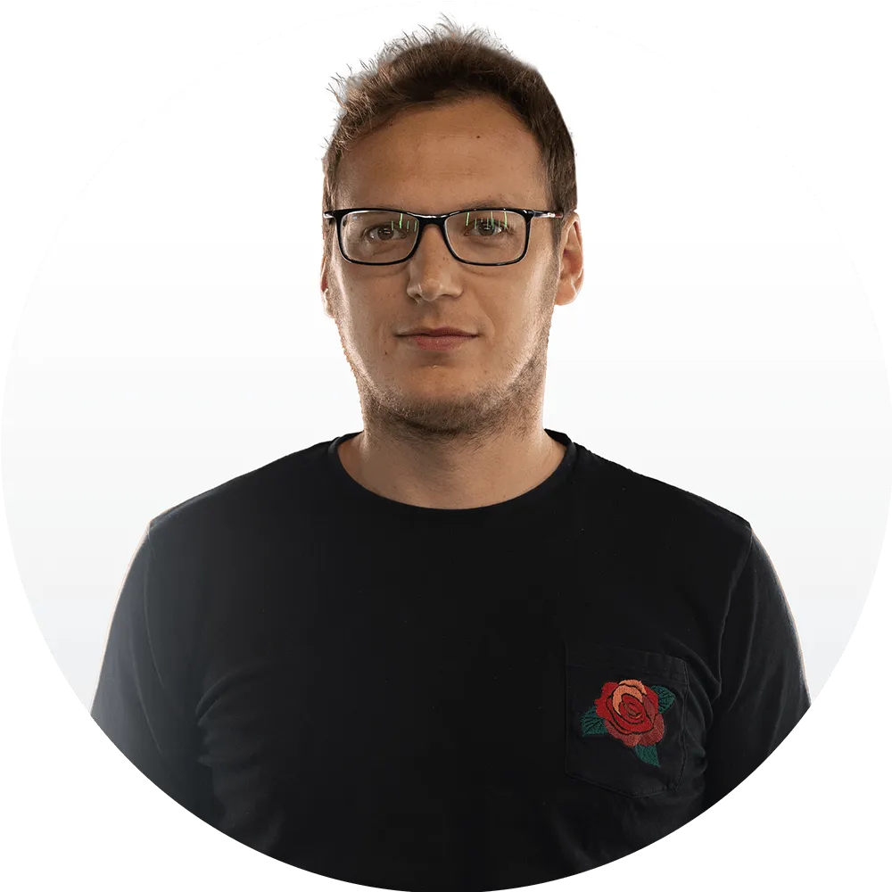 Piotr May A man with short brown hair and glasses wearing a black shirt with a red rose design on the pocket, poses against a plain white background.