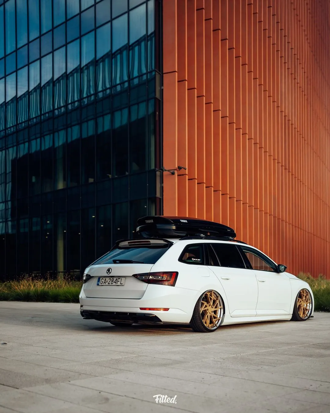 Ein weißer Kombi mit goldenen Felgen und einer Dachbox - gekennzeichnet als Projektbotschafter - steht neben einem modernen Gebäude aus Glas und orangefarbenen Paneelen, während das Sonnenlicht lange Schatten auf die Szene wirft.