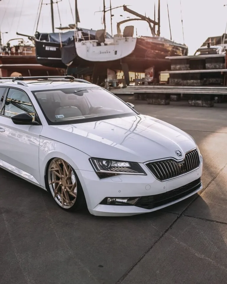 Ein weißer Skoda mit bronzenen Rädern, der als Projektbotschafter dient, ist auf einer Betonfläche in der Nähe mehrerer angedockter Segelboote und gestapelter Holzpaletten in einem Jachthafen geparkt.