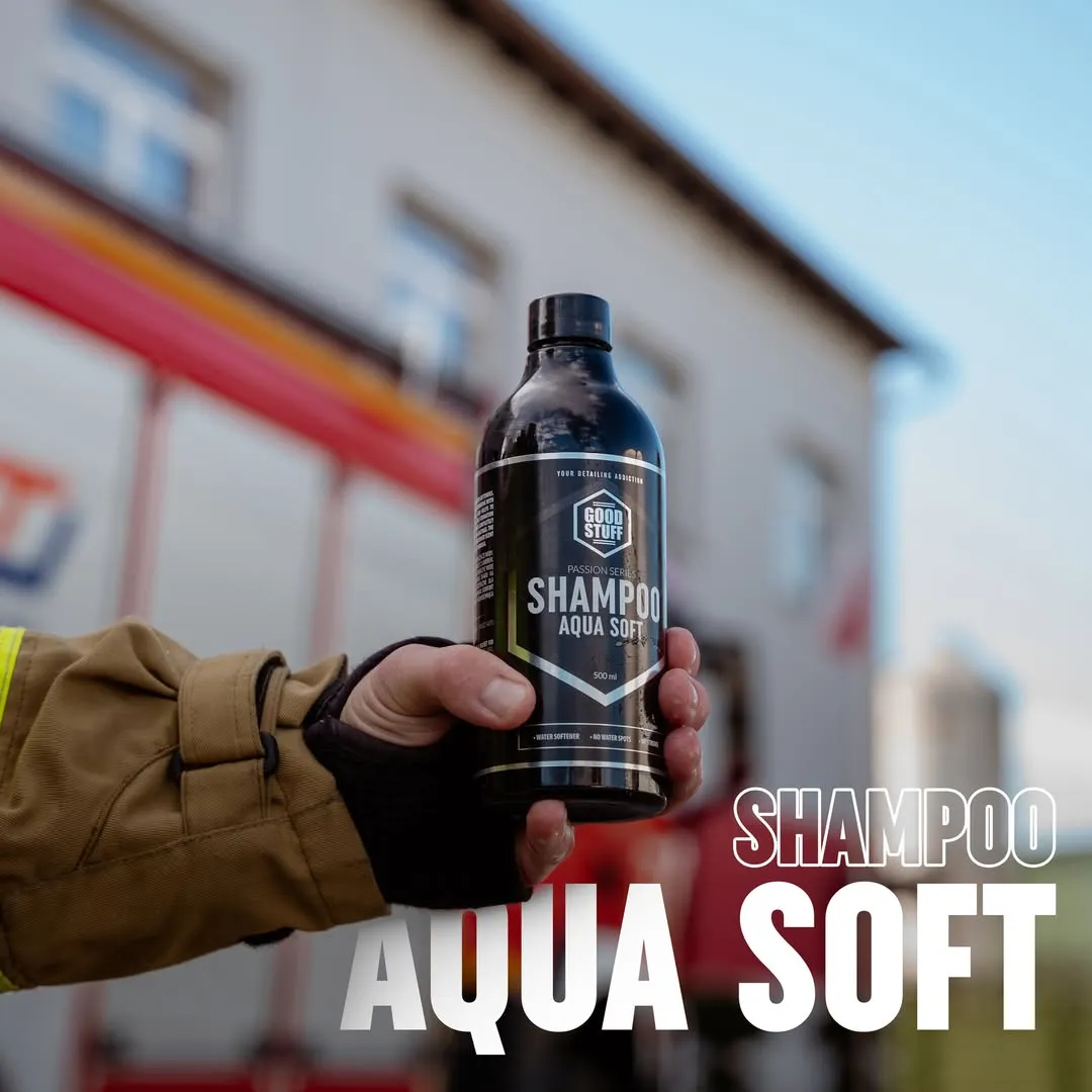 A person wearing a tan jacket and black glove holds a bottle of Good Stuff detailing shampoo outdoors, with a building and a red vehicle blurred in the background. Bold text reads SHAMPOO AQUA SOFT, highlighting this premium car wash product.
