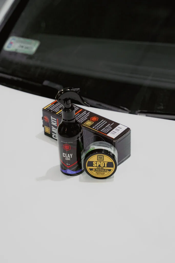 Autopflegeprodukte für die Lackdekontamination, darunter eine Clay Lube-Flasche und ein Spot-Behälter, stehen auf der Motorhaube eines weißen Fahrzeugs, dahinter befinden sich Verpackungen und im Hintergrund die Windschutzscheibe.