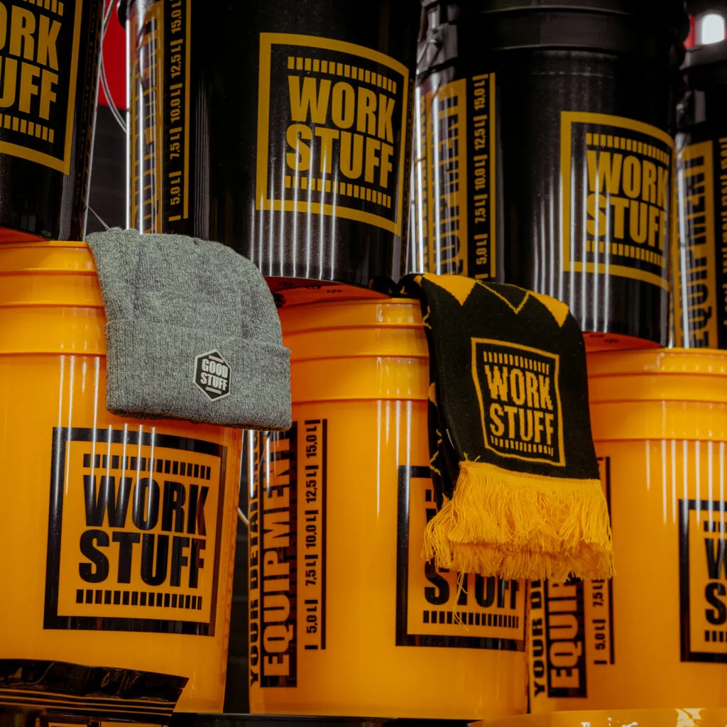 Una muestra de cubos WORK STUFF amarillos y negros apilados, con un gorro gris y una bufanda negra y amarilla sobre los cubos. El logotipo de la marca es visible en todos los artículos.