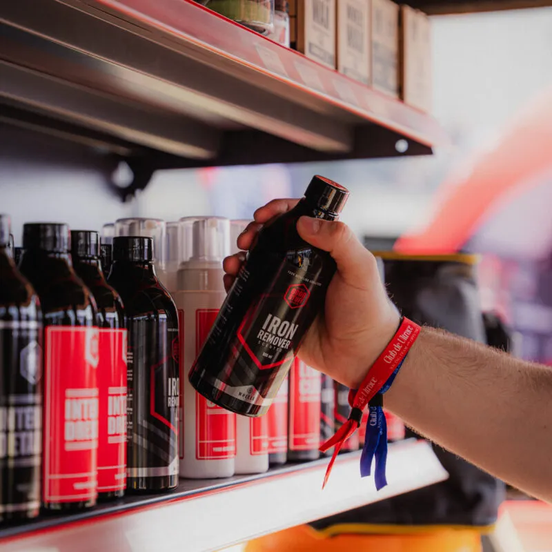 Osoba nosząca czerwoną i niebieską opaskę na nadgarstku trzyma butelkę z napisem Iron Remover na półce z różnymi produktami i butelkami do pielęgnacji samochodów.