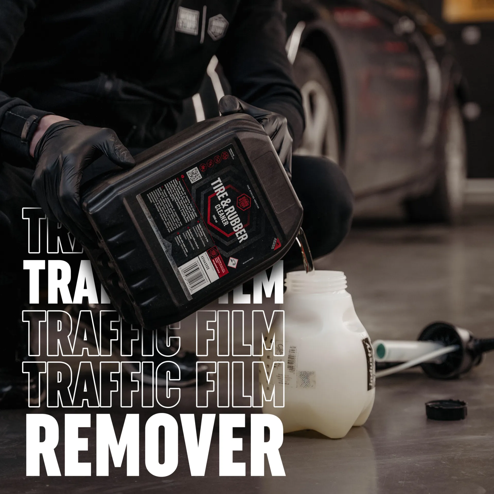 A person in black gloves pours Good Stuff Tire & Rubber Cleaner, a powerful pre-wash product and traffic film remover, into a white jug. A car is visible in the background with bold text reading “TRAFFIC FILM REMOVER.”.