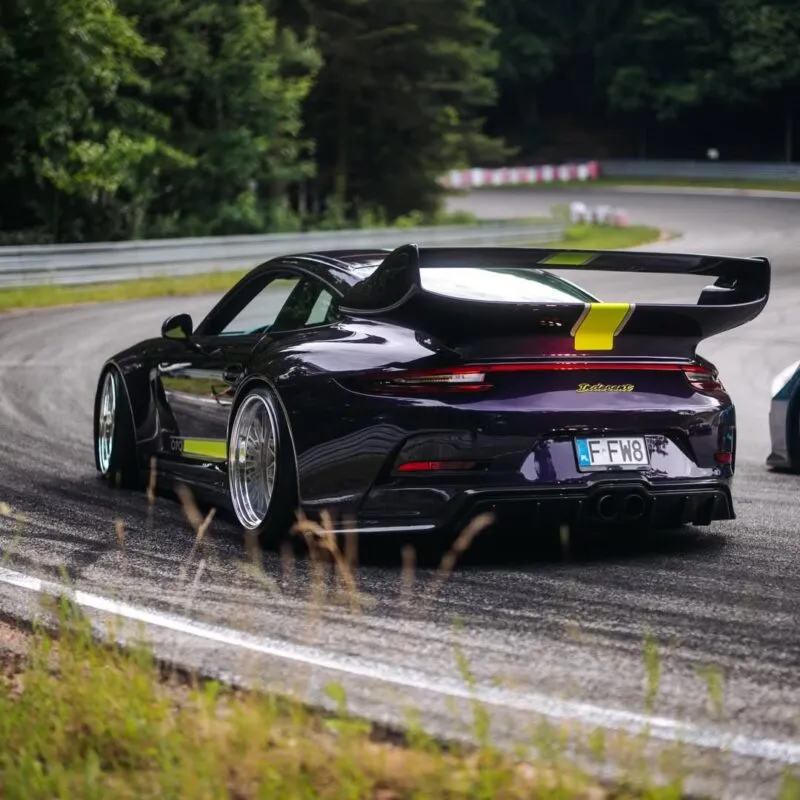 Un deportivo Porsche negro con un gran alerón trasero circula por una carretera de curvas rodeada de árboles y vegetación, acercándose a una curva de un circuito de carreras.