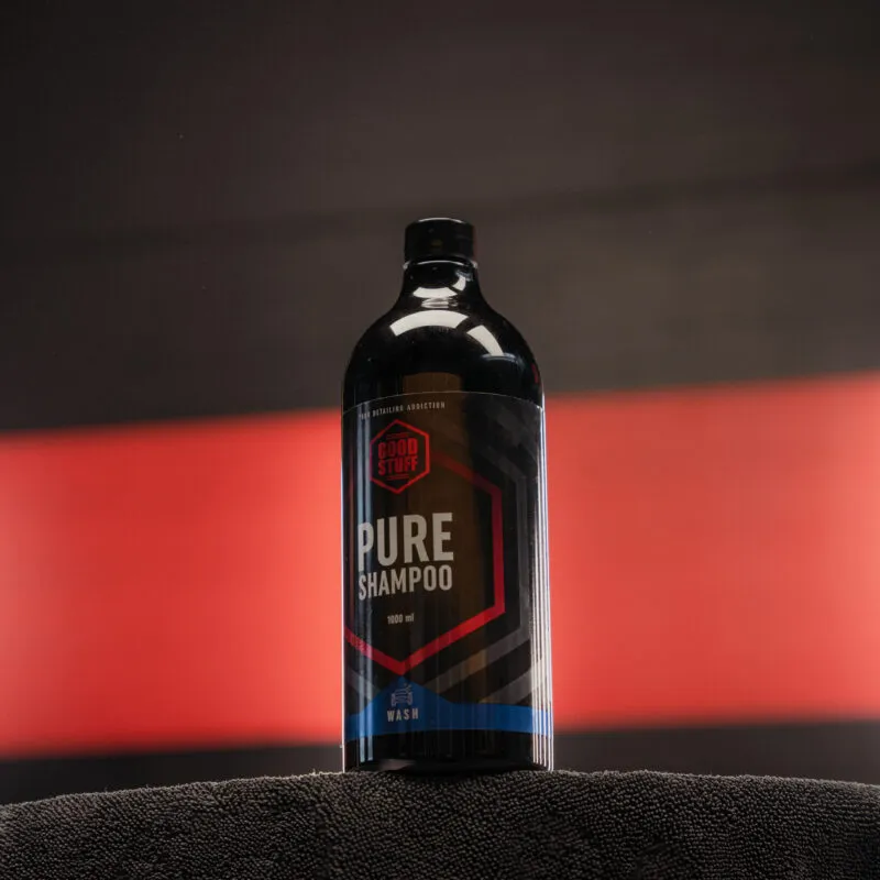 Czarna butelka szamponu samochodowego Pure Shampoo od Good Stuff stoi na ciemnym ręczniku z rozmytym czerwono-czarnym tłem. Etykieta ma sześciokątne kształty i pogrubione napisy.
