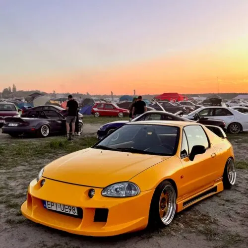 Ein leuchtend gelber, von Projektbotschaftern umgebauter Sportwagen ist auf einem Autotreffen auf dem Boden geparkt. Im Hintergrund sind bei Sonnenuntergang andere Autos, Menschen und Zelte zu sehen.