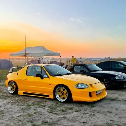 Ein leuchtend gelber, modifizierter Projektbotschafter-Sportwagen steht bei Sonnenuntergang auf einer Wiese bei einer Outdoor-Veranstaltung, mit Zelten, Menschen und einem weiteren geparkten Auto im Hintergrund.