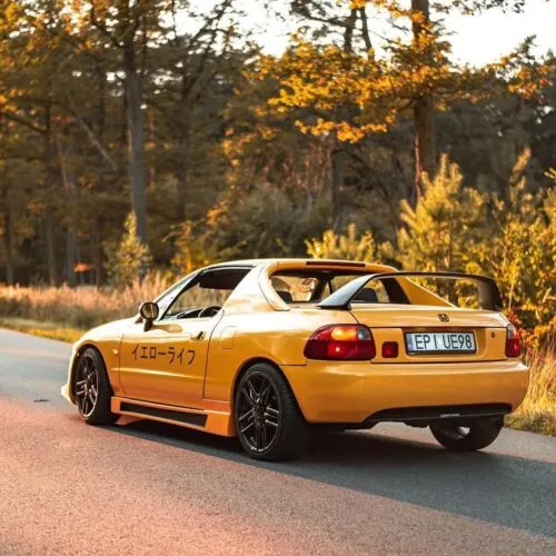 Ein leuchtend gelber Sportwagen mit schwarzen Felgen und einem Heckspoiler parkt auf einer von Bäumen gesäumten Straße im warmen, goldenen Sonnenlicht. Auf dem Nummernschild steht EP ULV89, japanische Schriftzeichen zieren die Seite und weisen ihn als echten Projektbotschafter aus.