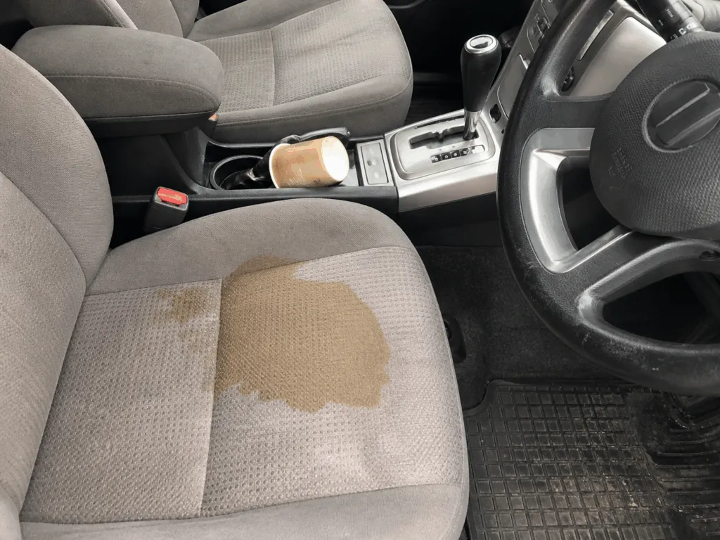 A spilled coffee cup lies on the center console of a car, with a large coffee stain soaking into the gray fabric of the front passenger seat—a perfect moment to consult a car shampoo guide for tackling stubborn stains and keeping your car interior spotless.