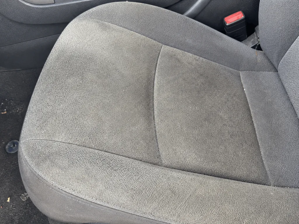 A close-up view of a car seat with dark gray upholstery, showing some slight wear and a seat belt buckle to the upper right. The clean and well-maintained area suggests regular care—perhaps following a car shampoo guide for best results.