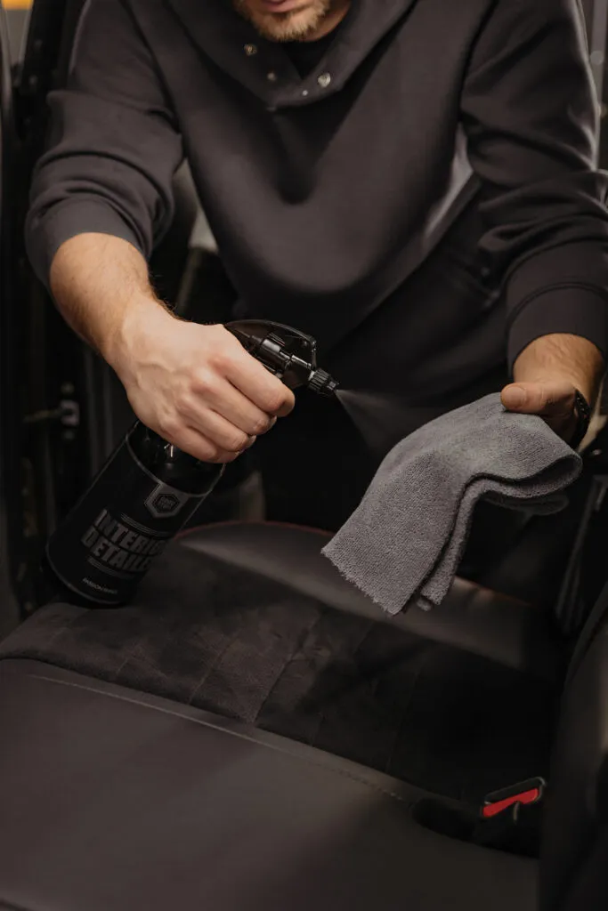 A person is spraying cleaner from a black bottle onto a gray microfiber cloth while cleaning the interior of a car seat, ensuring clean interior fabric—perfect for families with kids and pets or tackling coffee stain removal.