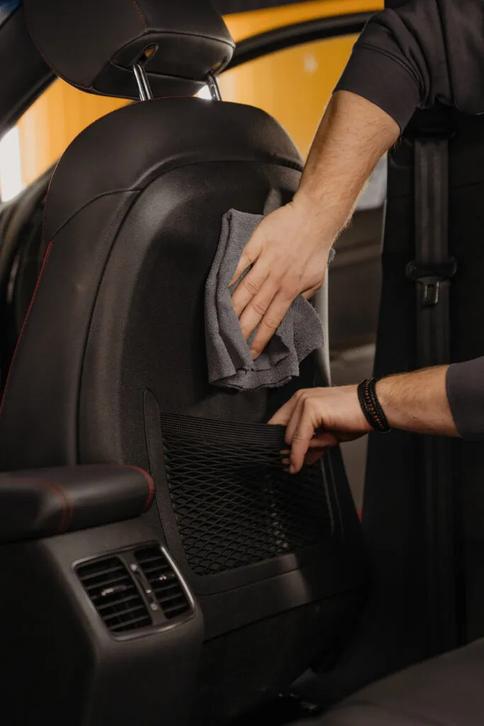 A person cleaning the back of a car seat with a gray cloth, focusing on clean interior fabric while holding a black storage pocket. The interior of the car is visible in the background.