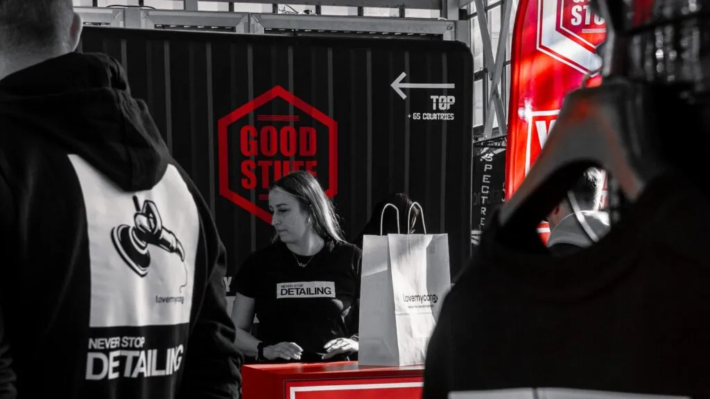 Una mujer detrás de un mostrador rojo con bolsas de la compra en un puesto de limpieza de coches. Lleva una camiseta de Never Stop Detailing. Detrás de ella hay un cartel rojo en el que se lee GOOD STUFF con una flecha encima. Otras personas son parcialmente visibles.
