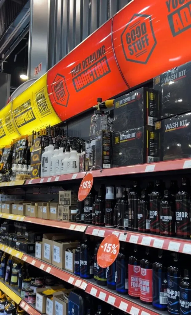 En los estantes de una tienda de limpieza de coches se exponen productos de limpieza, como botellas, sprays y kits. Sobre los estantes cuelga un gran kayak rojo con texto publicitario, y se ven varios carteles rojos de -15% de descuento.