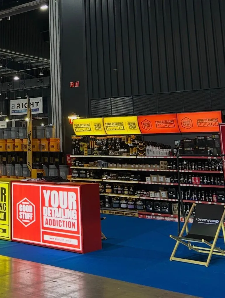 En un stand de feria se exponen productos para el cuidado del automóvil en estanterías con carteles luminosos en los que se lee GOOD STUFF y YOUR DETAILING ADDICTION. Cerca hay dos sillas plegables sobre una alfombra azul.