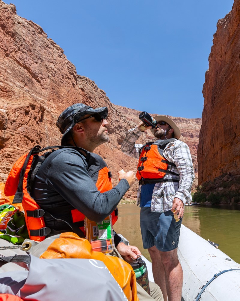 Rafting with Hatch River Expeditions two men wearing life vests on a river raft in Grand Canyon who are looking at the cliffs while drinking water