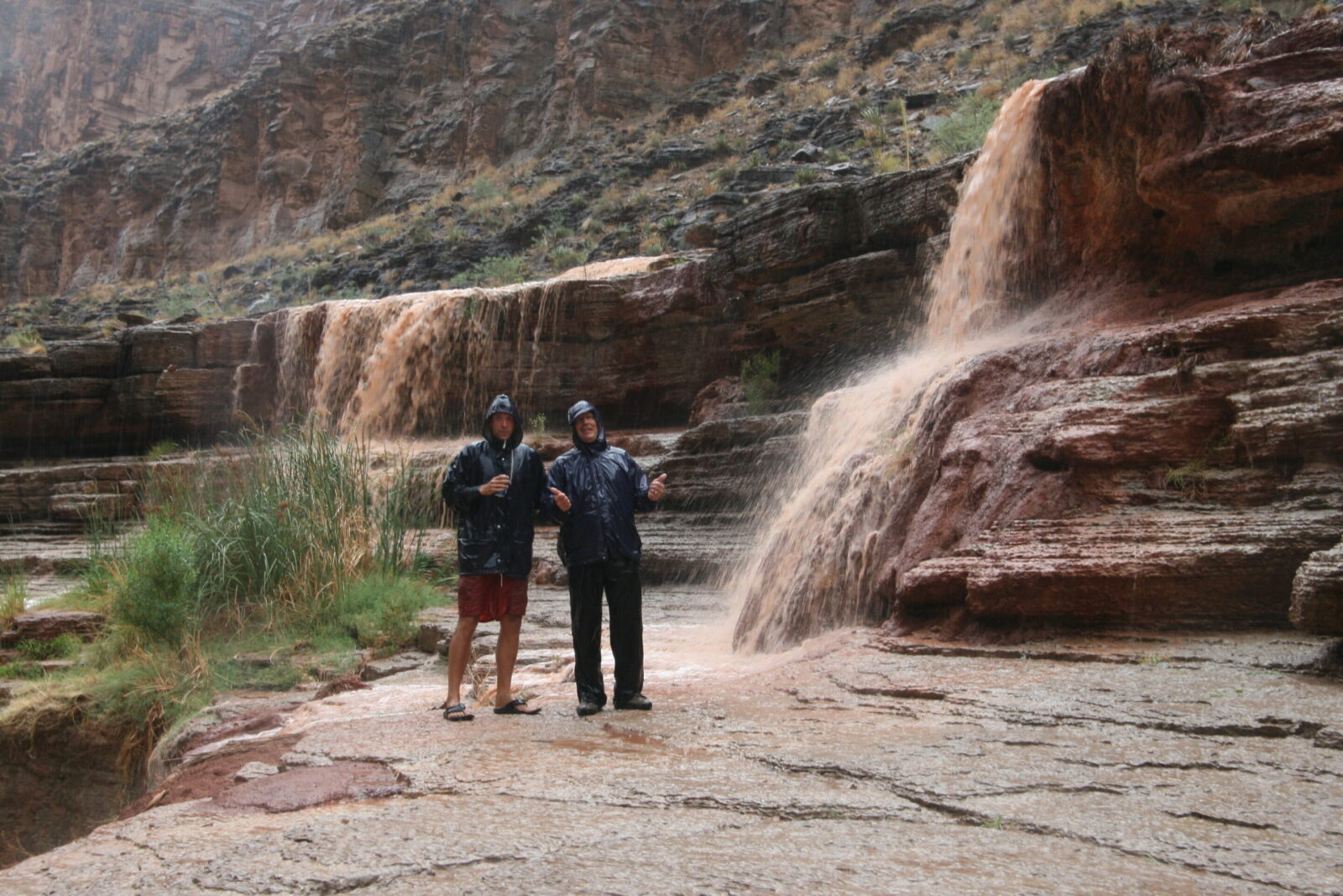 Raining near waterfalls of Grand Canyon with Hatch River Expeditions Two men in rain gear stand in the rain in Grand Canyon near an ephemeral waterfall pouring down the canyon cliffs. Blog on tips for if it rains on your rafting trip.