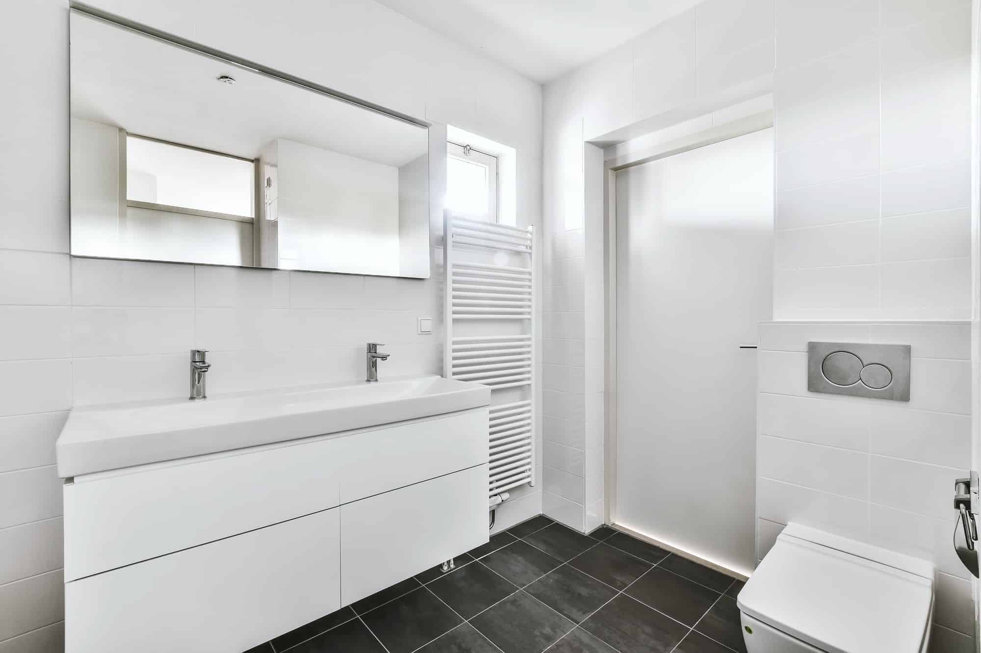 KL Plomberie Services Intérieur de salle de bains moderne avec murs blancs, vanité flottante, grand miroir, porte-serviettes chauffant et sol carrelé foncé.