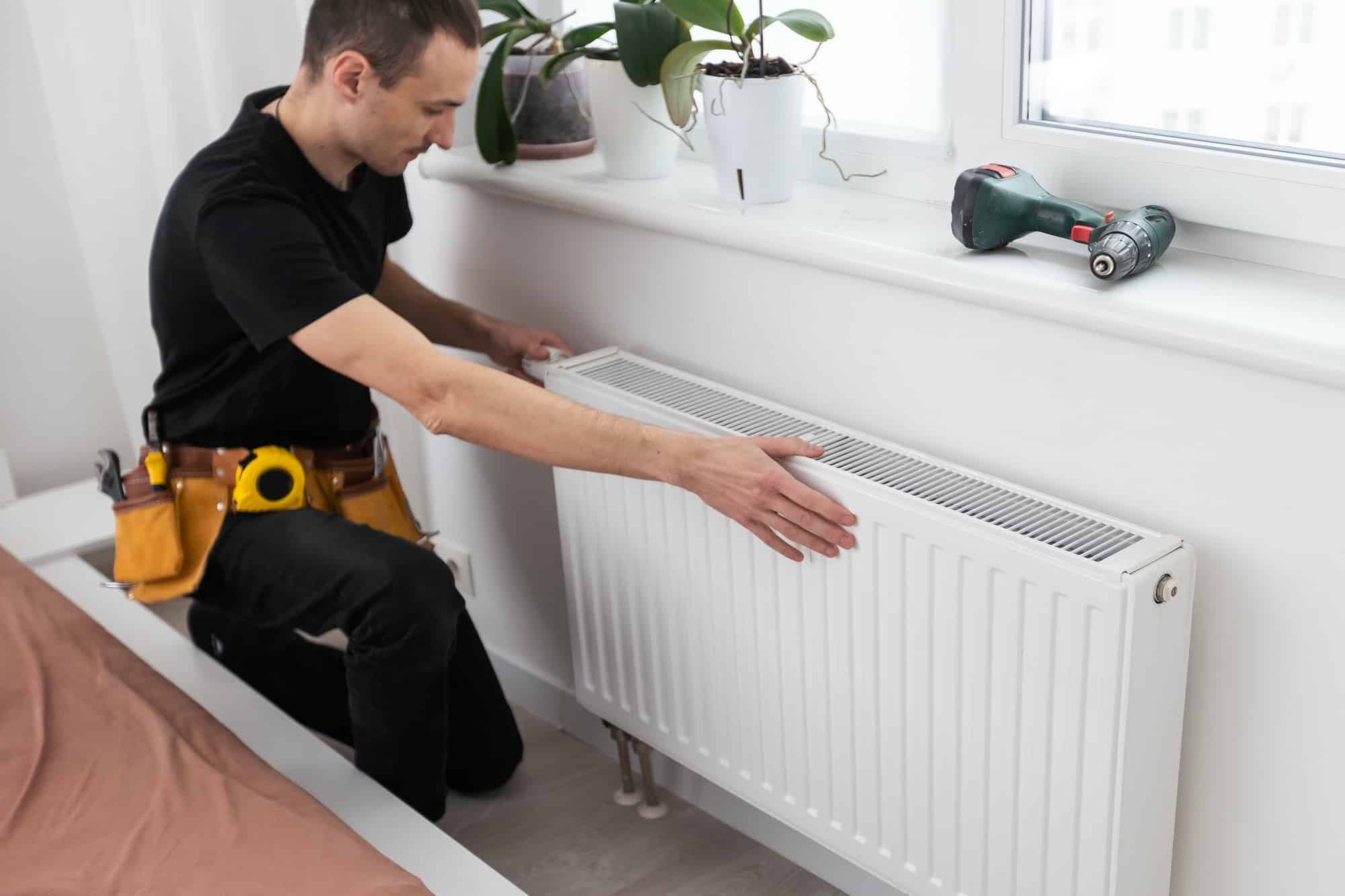 KL Plomberie Services Un homme muni d'une ceinture à outils installe un radiateur sous une fenêtre, avec une perceuse sans fil à proximité.