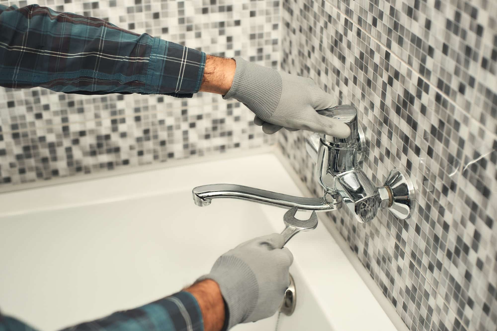 KL Plomberie Services Une personne portant des gants ajuste un robinet moderne au-dessus d’une baignoire blanche, contre un mur carrelé.