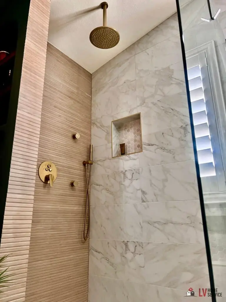 Modern shower with a rain showerhead, gold fixtures, marble tiles, and a recessed shelf.