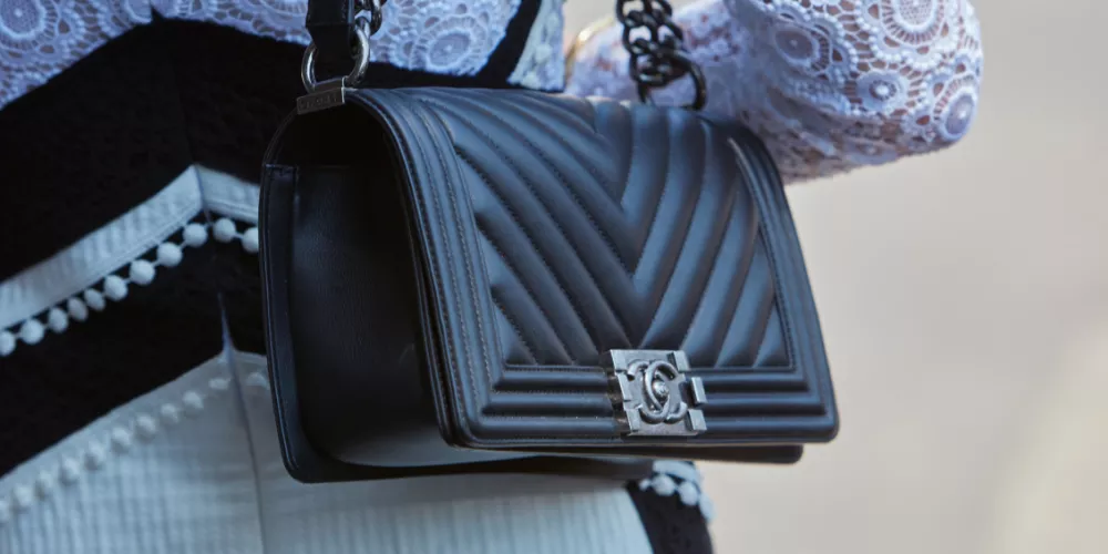 Close Up Of A Black Quilted Leather Handbag With A Chain Strap And Metallic Clasp, Worn Over A White Lace Sleeve And Black And White Outfit, Beautifully Showcasing The Elegance Associated With Exploring Chanel's Leather Varieties.