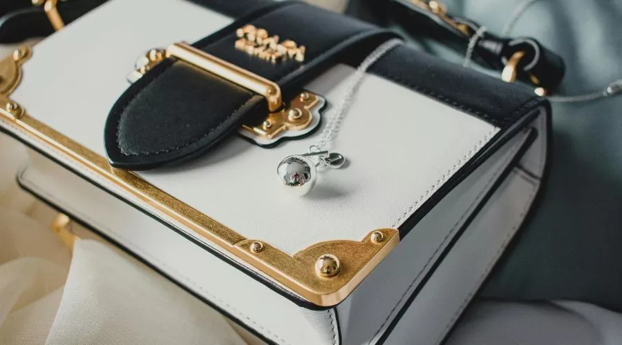 A Black And White Handbag Crafted From Top Grain Leather, With Gold Accents, Is Placed On A Fabric Surface. A Small Silver Ball Pendant Necklace Rests Elegantly Atop The Bag.