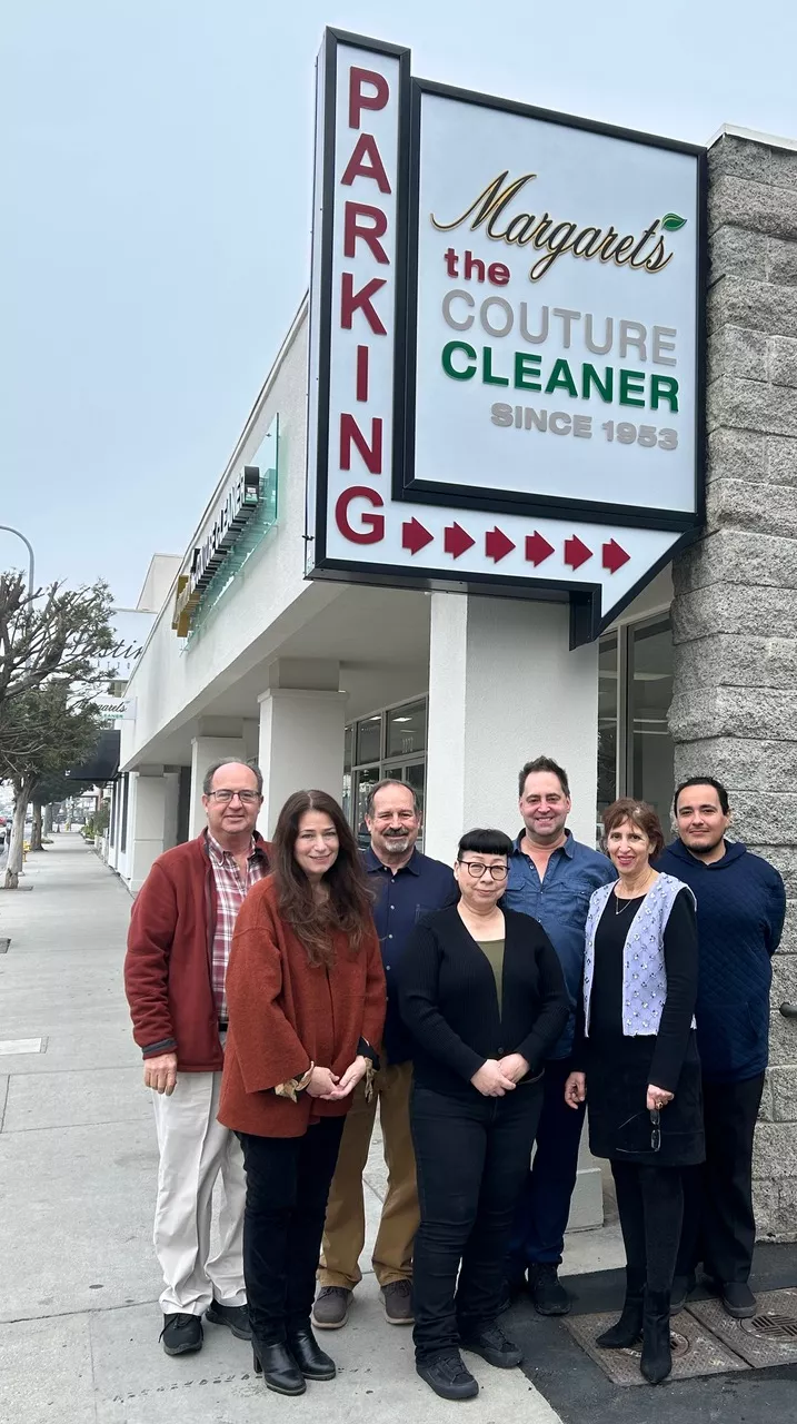 Group Of Seven People Standing In Front Of Margaret's The Couture Cleaner Sign Outside A Building.