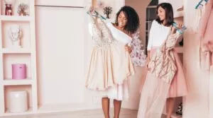 Two Women In A Boutique, Smiling And Holding Up Formal Dresses Ready For Dry Cleaning. One Dress Is Cream With Embellishments; The Other Is Pink With Patterns. Shelves And A Rack Of Clothing Are In The Background.